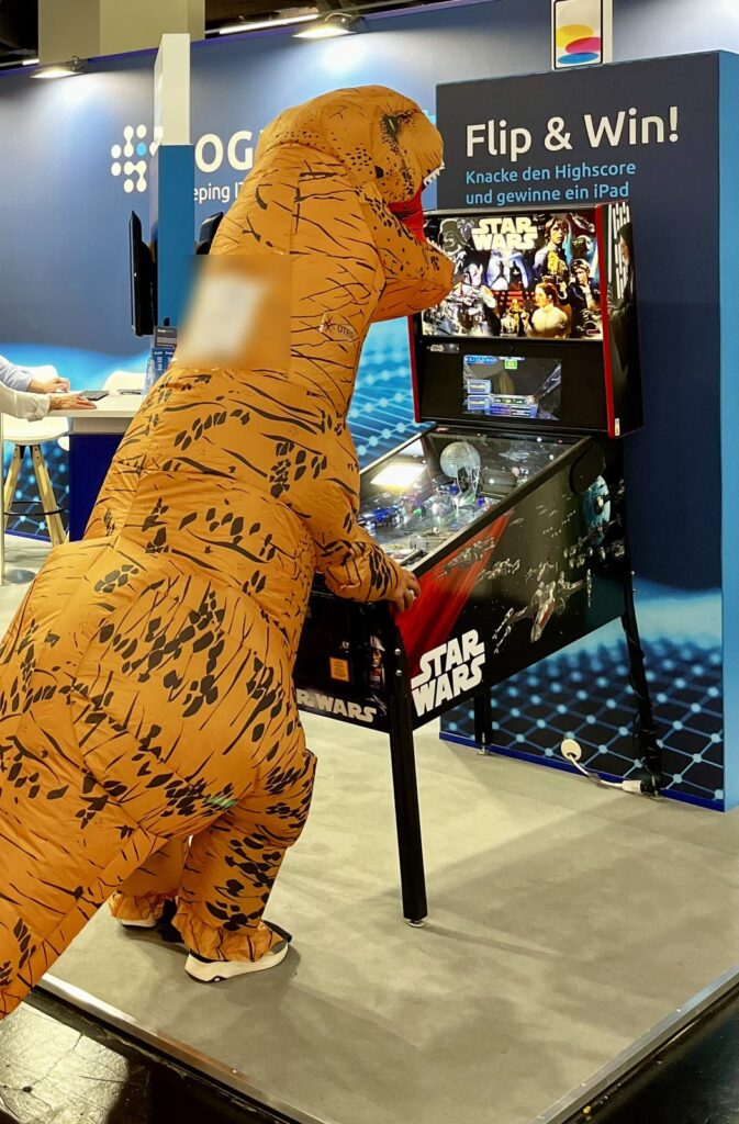 Person im aufblasbaren T-Rex-Kostüm spielt am Star-Wars-Flipper auf dem LOGINventory-Messestand der it-sa 2024